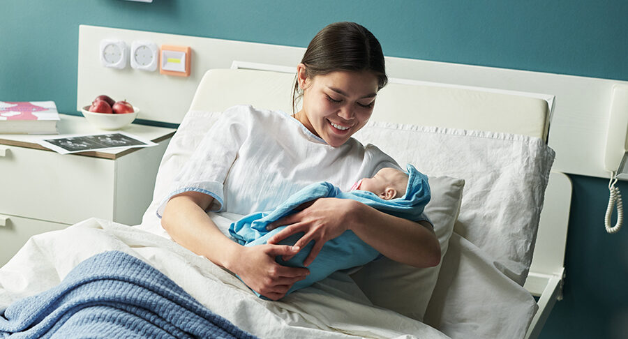 Young Caucasian woman lying in hospital bed holding newborn baby, smiling and looking at infant,